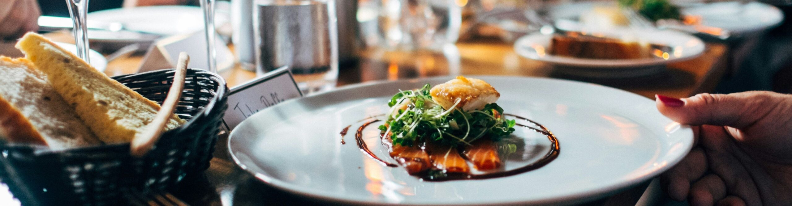 Food on a plate in a restaurant with someone sat at the table. Bread and glasses on the table